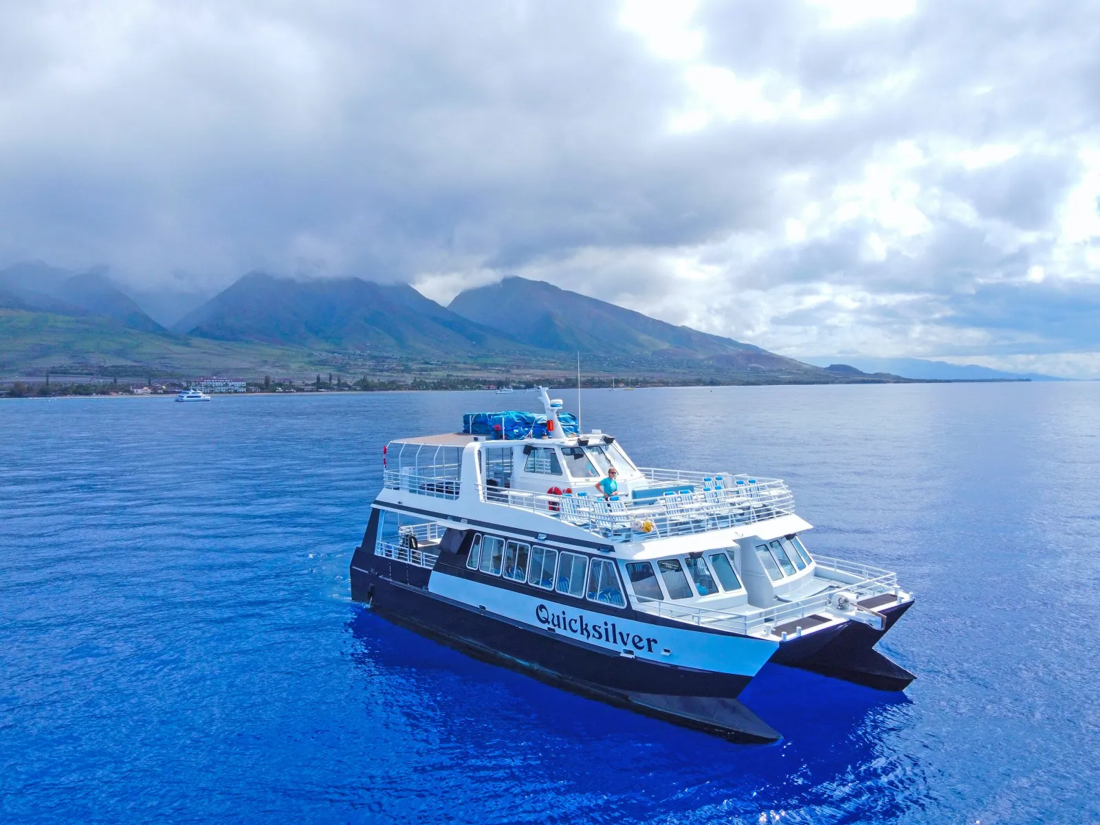 A white double-deck power catamaran named Quicksilver on blue water below cloud-topped mountains