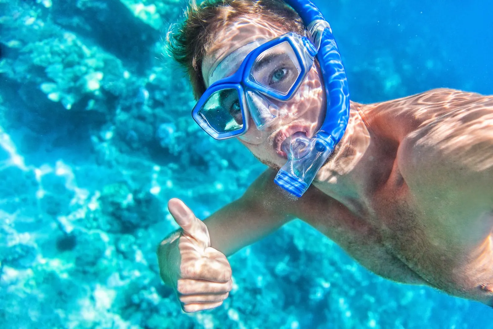 Smiling snorkeler in a blue mask giving a thumbs-up underwater over coral