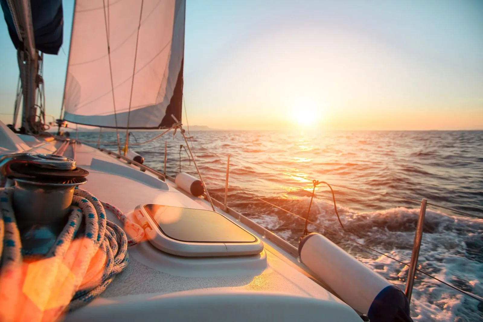 Sailboat deck with winch and coiled rope heeling toward a low, bright sun