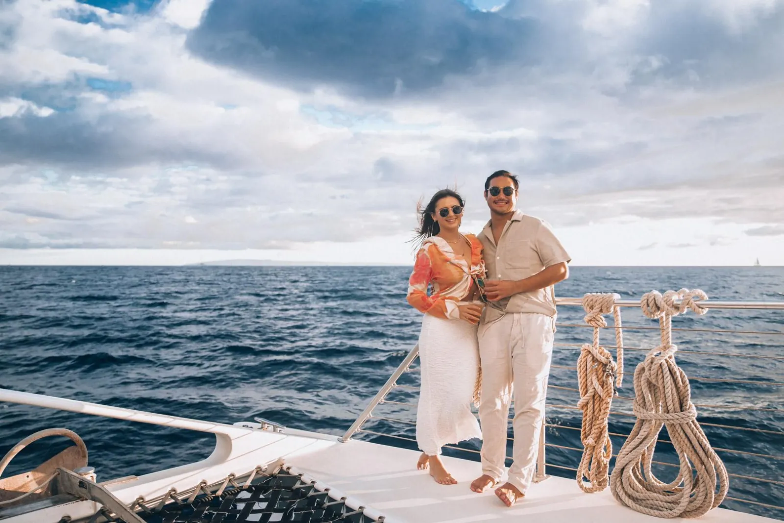 Barefoot couple holding drinks at a catamaran's bow rail under clouds