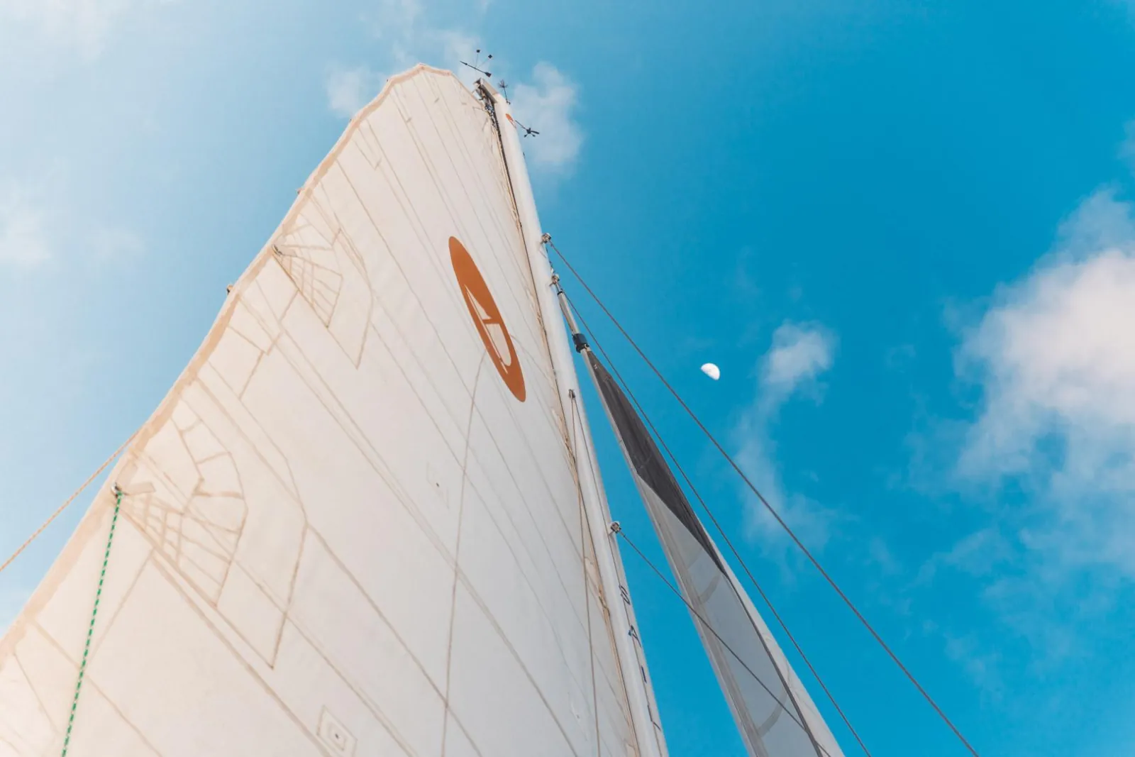 Looking up at a white mainsail against blue sky with a daytime moon