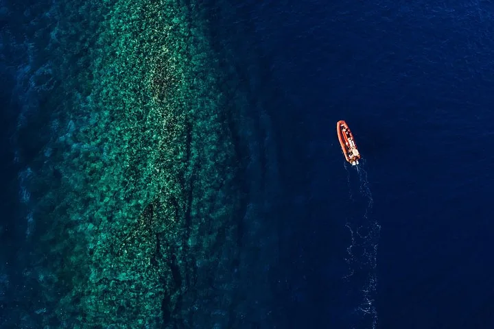 Aerial view of an orange raft passing a shallow turquoise reef in dark blue water