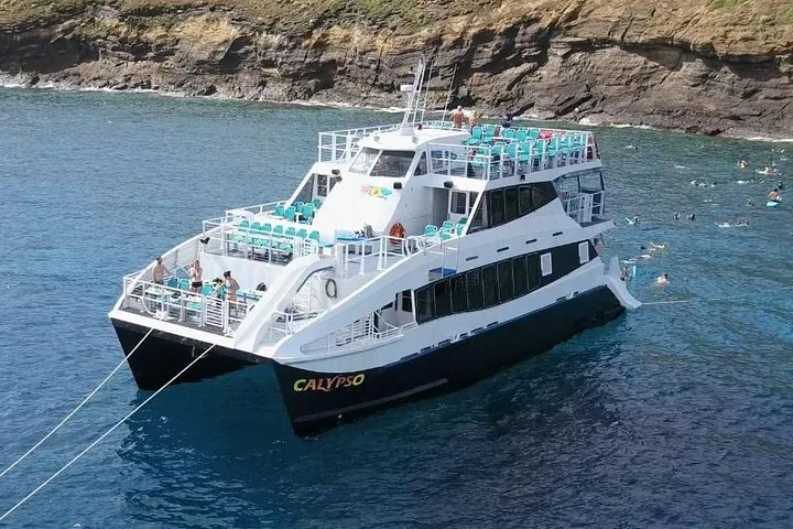 White three-deck Calypso catamaran moored by a rocky cliff with snorkelers in the water