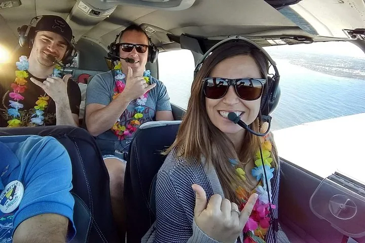 Three passengers in headsets and lei giving shaka signs inside a small plane cabin