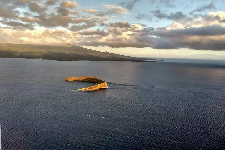 Crescent-shaped Molokini seen from the air with a mountainous coastline and clouds behind